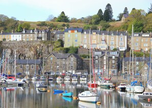 Un port où sont amarrés de nombreux bateaux, des bâtiments en pierre à plusieurs étages et une colline parsemée d’arbres, avec une maison visible au loin.