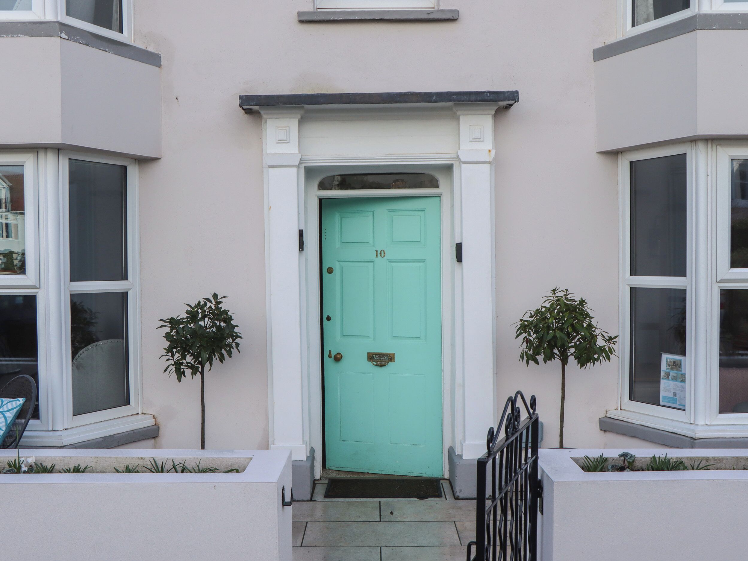 A turquoise door with a white frame, flanked by two small potted plants, leading to a white entrance with a black metal gate.