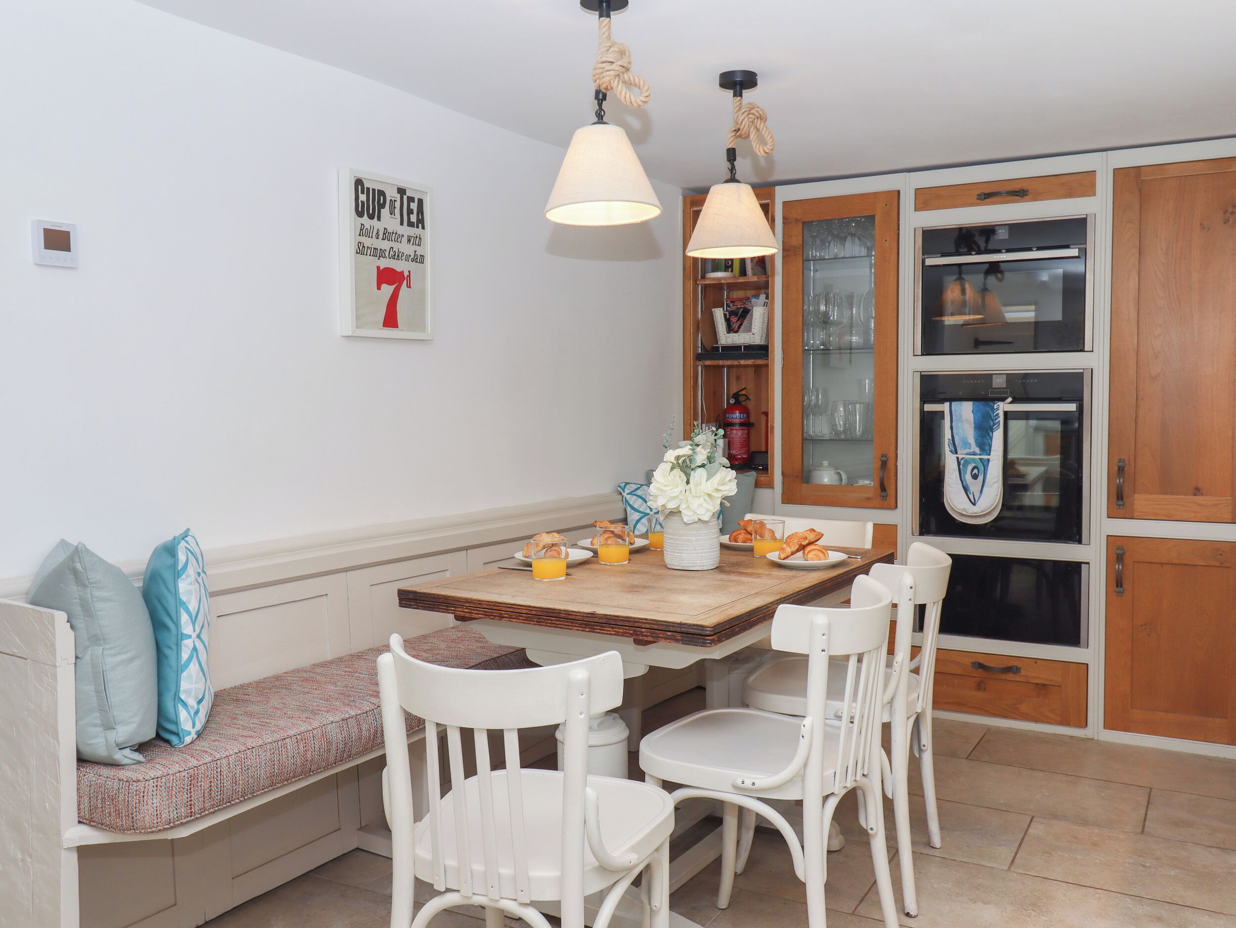 A dining area with a wooden table, white chairs, a bench with cushions, a hanging light fixture, and a kitchen area with cabinets and a stove.
