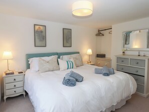 A neatly made bed with white bedding and striped pillows, a bedside table with a lamp, a dresser with drawers, and a mirror on the wall. - Sanctuary Cottage (Teignmouth)