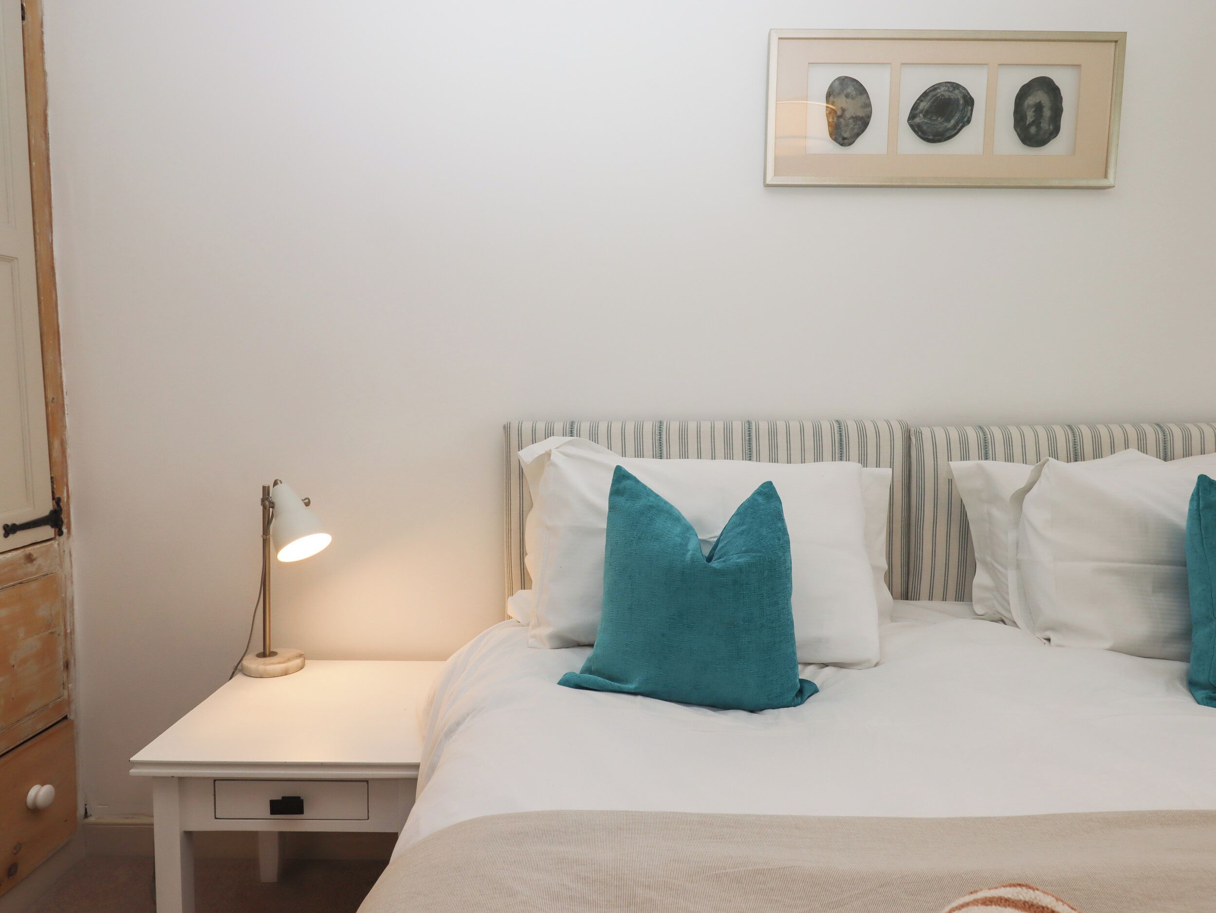 A neatly made bed with white bedding and teal pillows, a bedside lamp, and a framed picture above the bed.
