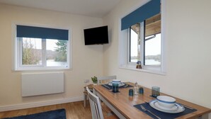A compact kitchen with a wooden table, two windows with blue blinds, a mounted TV, and a view of a lake. - Kingfisher Lodge (St Austell)