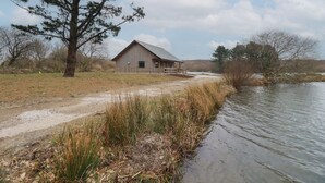 A rustic wooden cabin by a lake with a dirt path leading to it. - Kingfisher Lodge (St Austell)