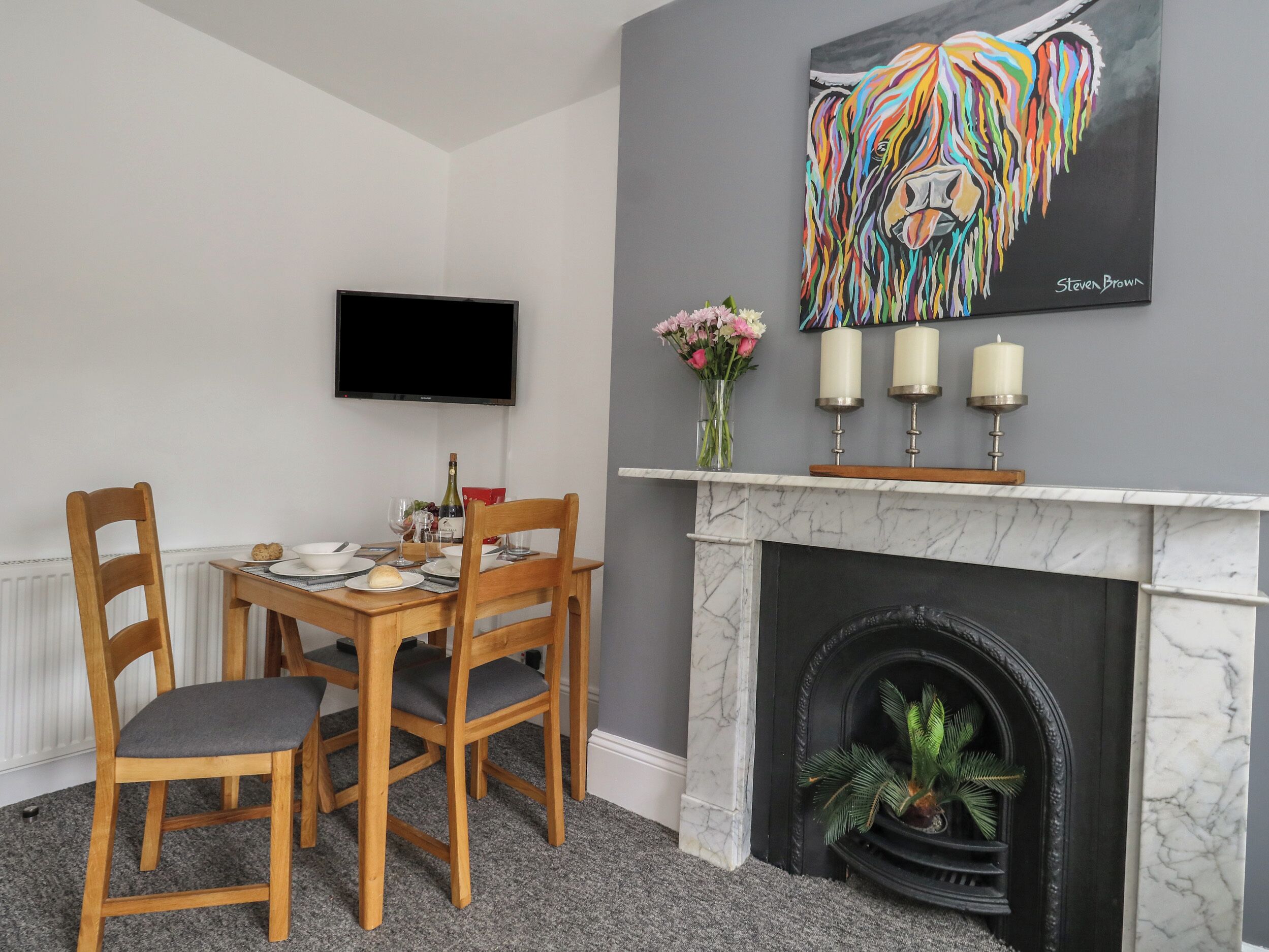 A dining area with a wooden table and chairs, a flat-screen TV mounted on the wall, a fireplace with a marble surround, and a colorful lion painting above the mantel.