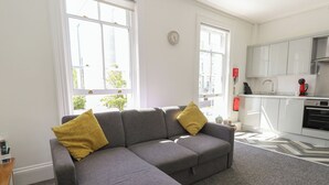 A modern living room with a grey sectional sofa, two yellow cushions, a grey carpet, and a kitchen area with white cabinets and appliances. - Flat 3 - Muderford (Bournemouth)