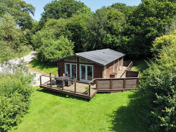 A wooden cabin with a deck, surrounded by lush greenery. - 1 Hedgerows (Looe)