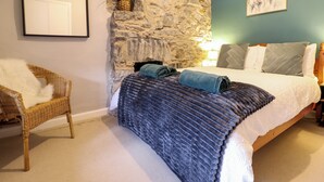 Een slaapkamer met een stenen muur, een bed met een blauwe deken, een rieten stoel en een ingelijst beeld aan de wand.