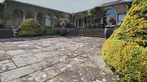 A courtyard with stone flooring, a large bush, and a building with a tiled roof and columns.