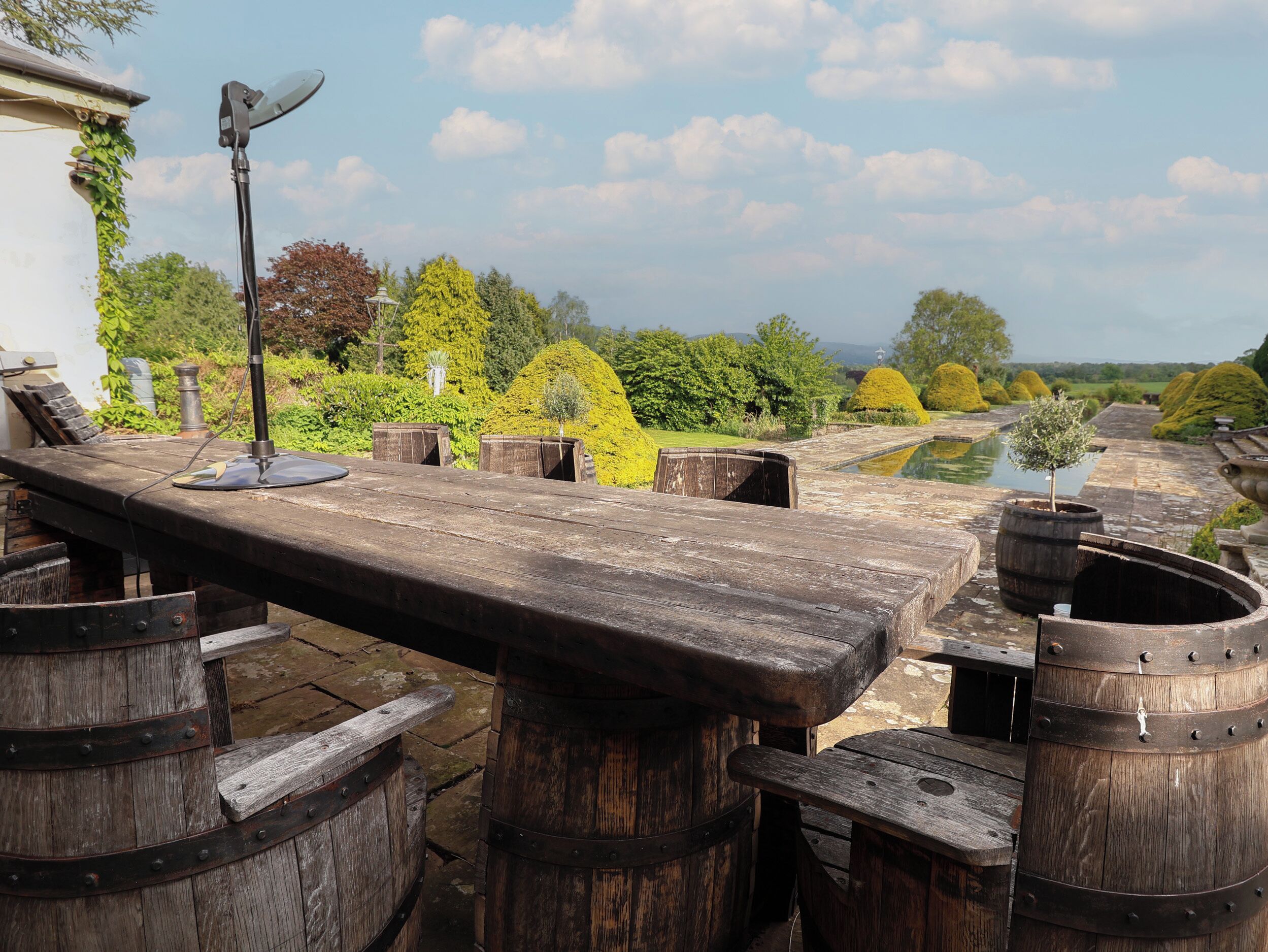 A rustic outdoor seating area with wooden tables and barrels, surrounded by lush greenery and a clear sky.