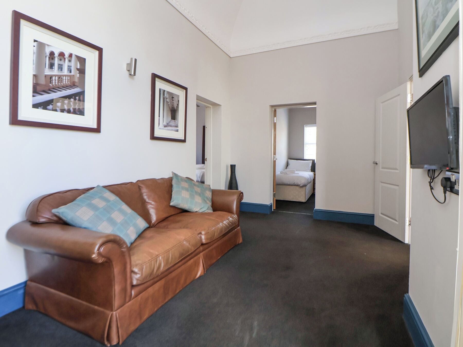 A hotel room with a brown leather sofa, two framed pictures on the wall, a television, and a door leading to another room.