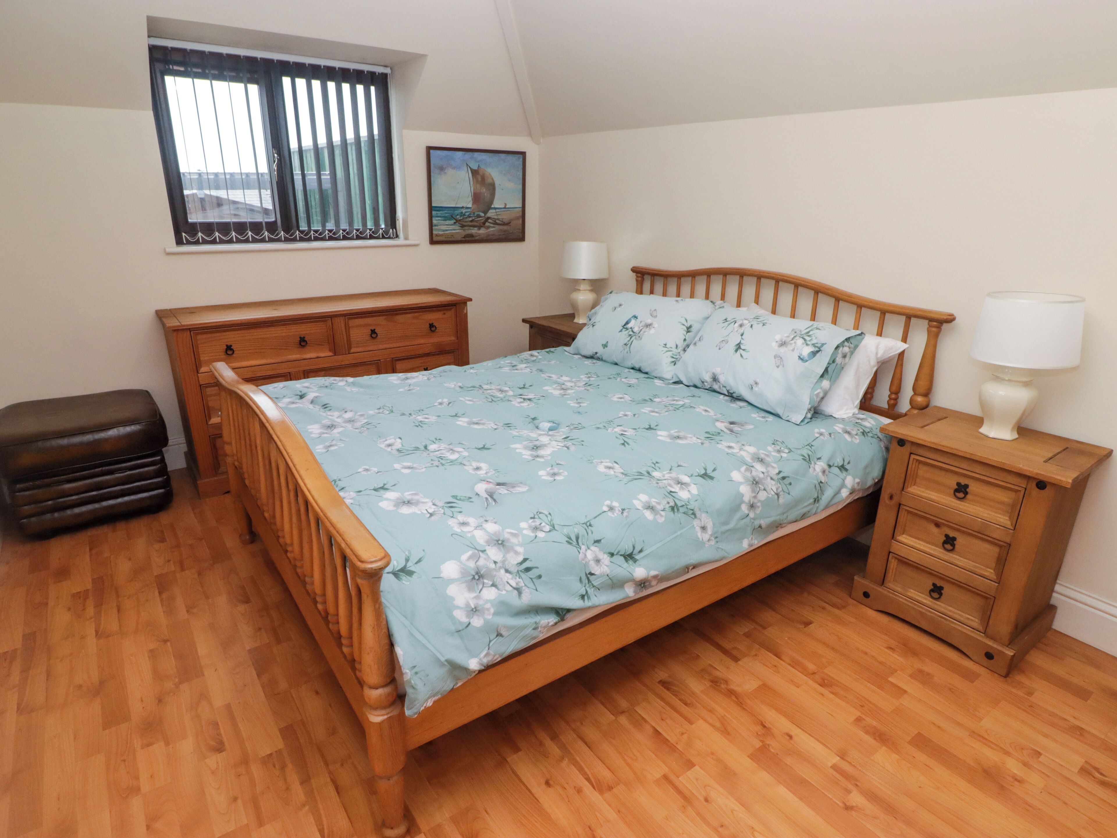 Une chambre avec un lit en bois, une table de chevet avec une lampe, un banc en bois, une fenêtre avec des stores et un tableau accroché au mur.