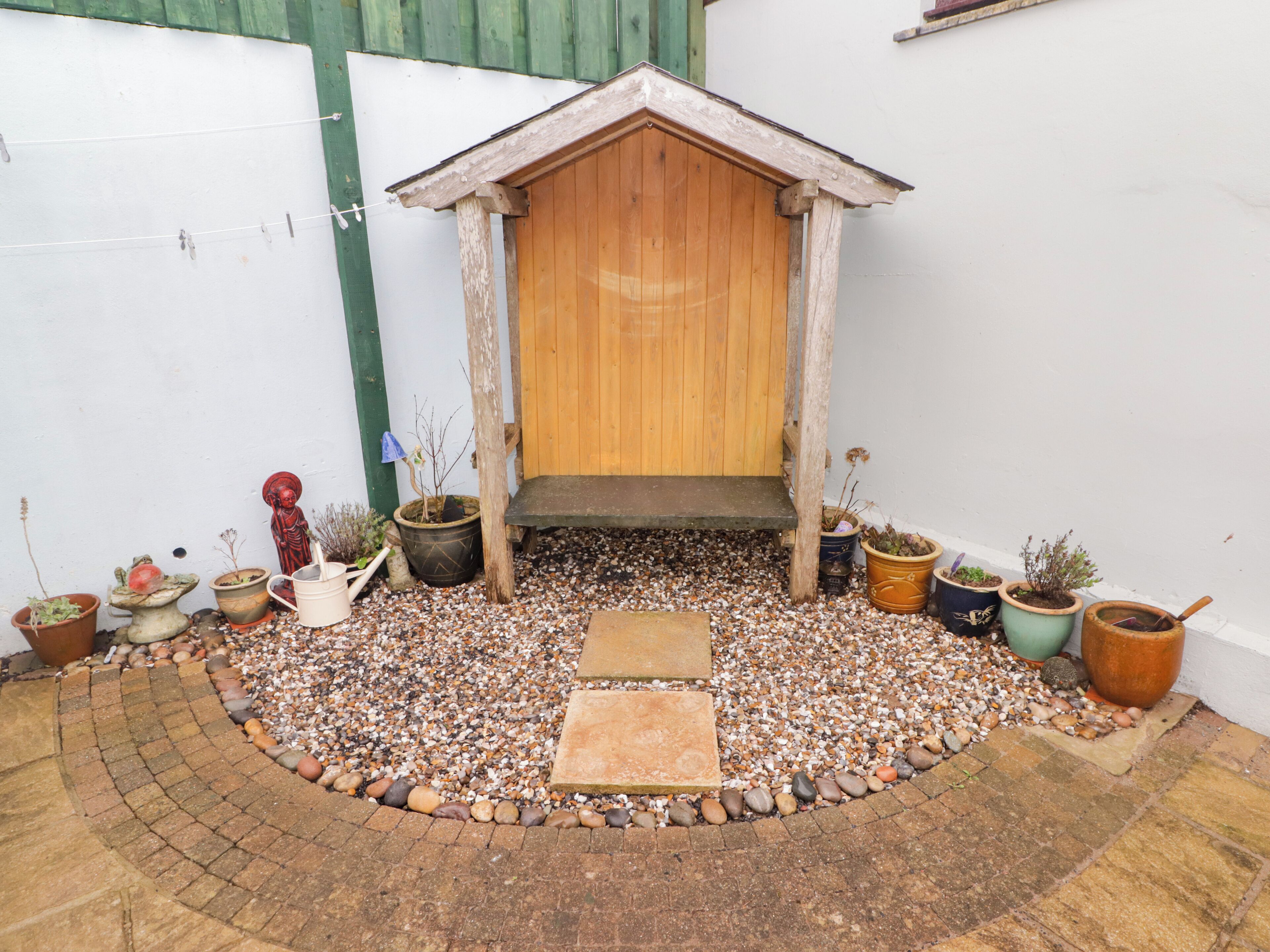 Une petite construction en bois, avec un banc devant, entourée d’un chemin de gravier et de différents pots de plantes.