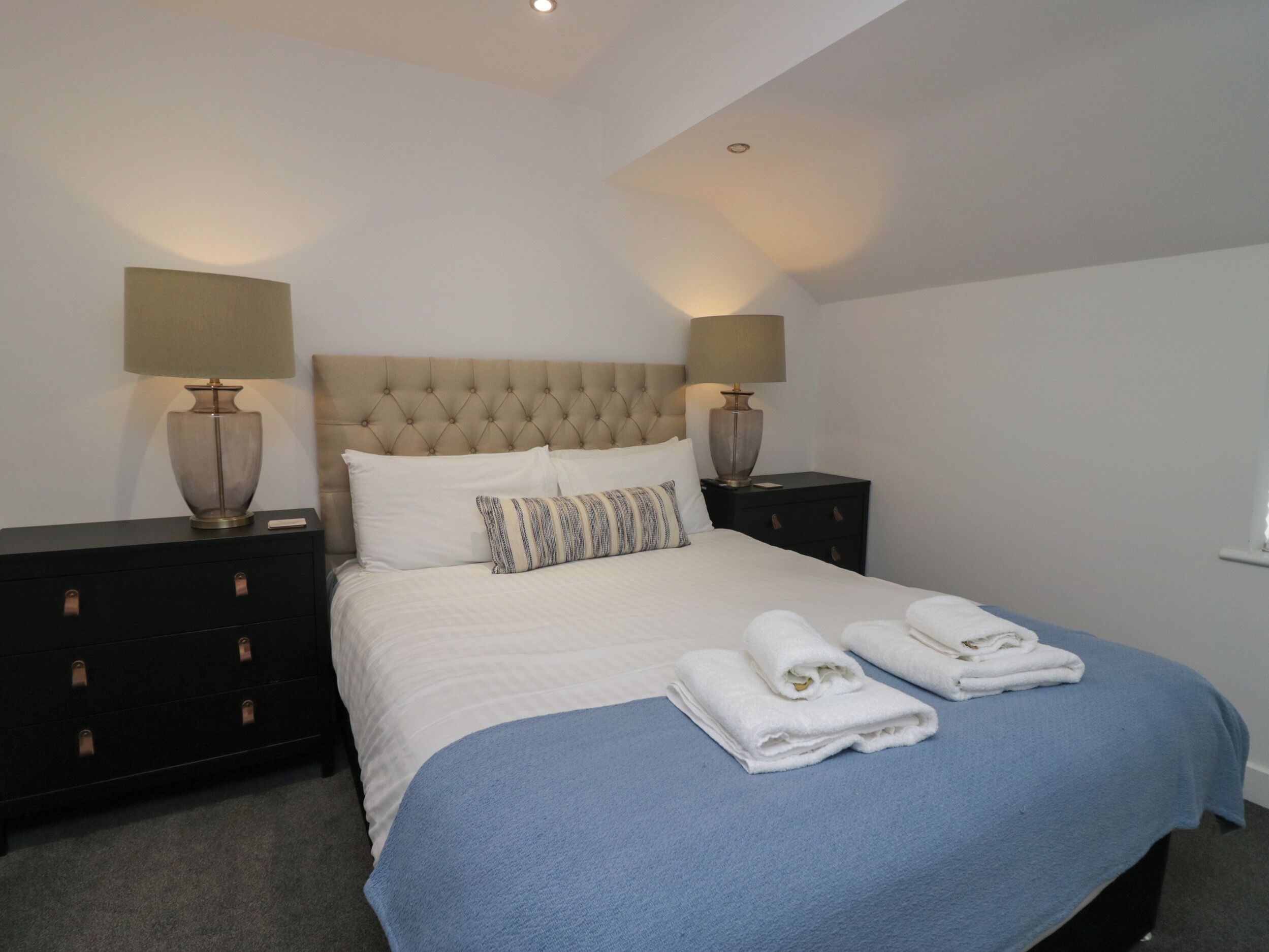 A neatly made bed with white linens and a blue blanket, flanked by two bedside tables with lamps, in a simple room with white walls and a tufted headboard.