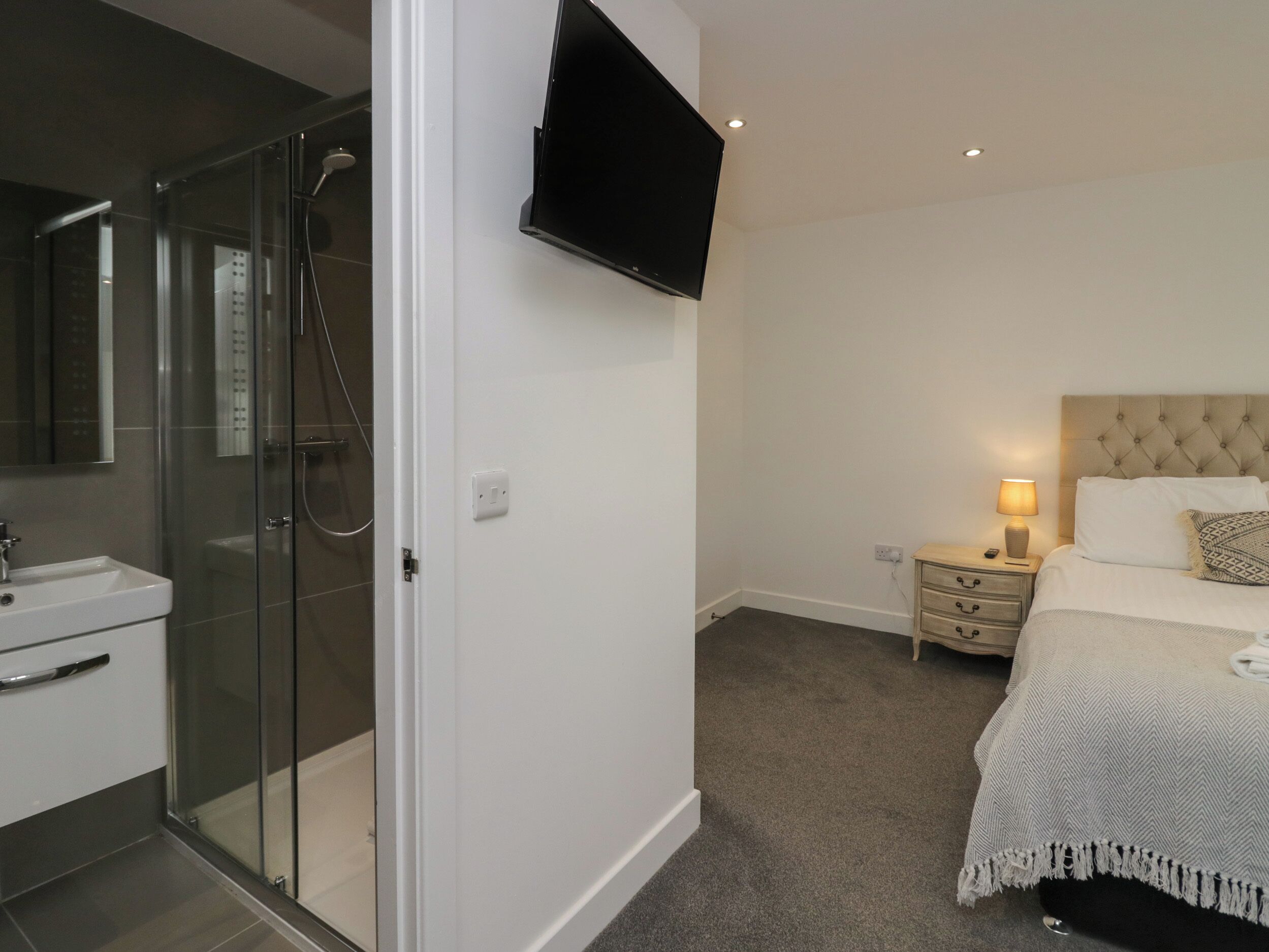A modern bedroom with a glass-enclosed shower, a flat-screen TV mounted on the wall, a bedside table with a lamp, and a bed with a patterned bedspread.