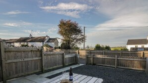 A wooden picnic table with a bottle and hats, surrounded by a wooden fence and houses in the background. - Edgemere Hideaway (Grange-over-Sands)