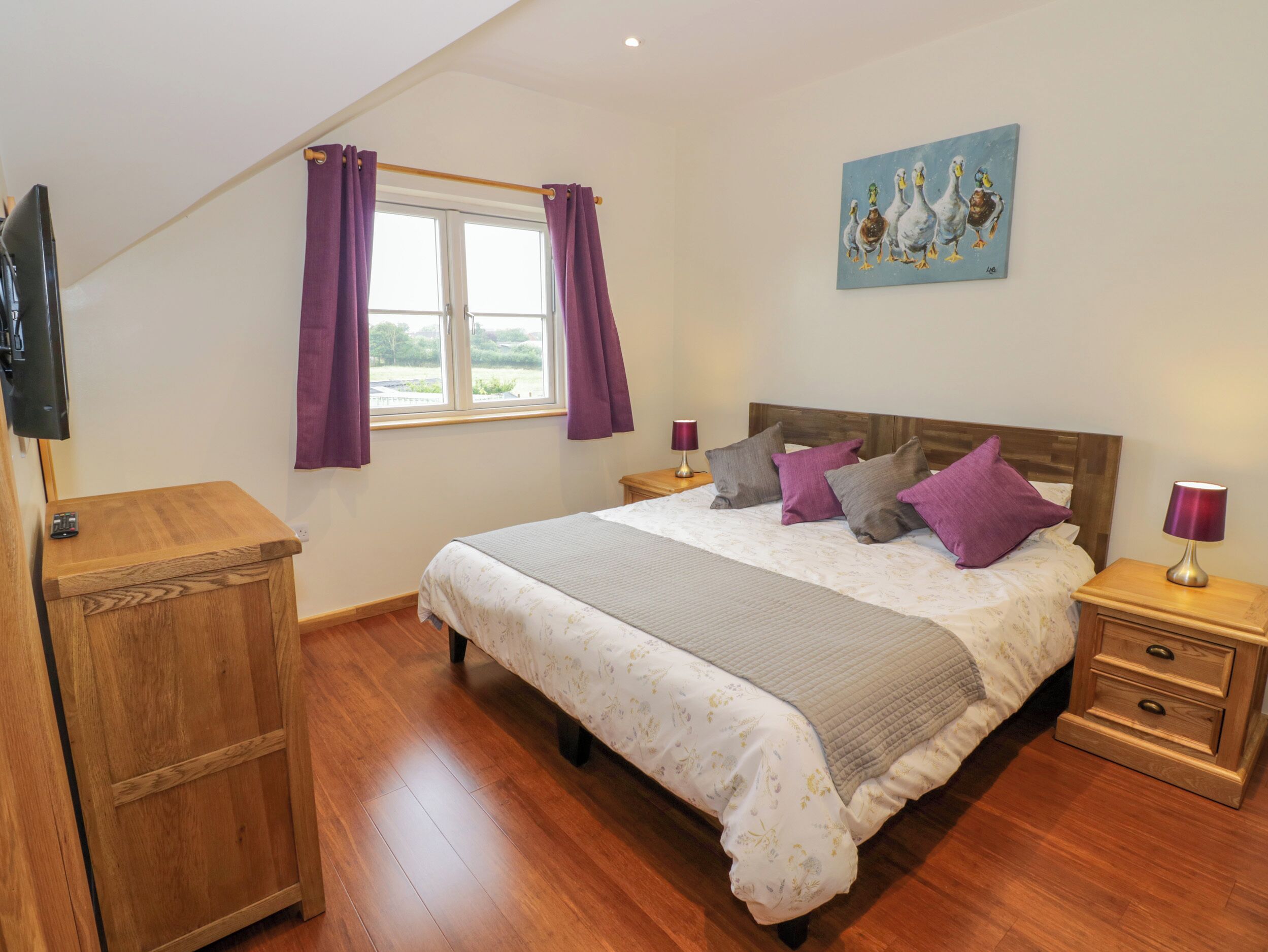 A bedroom with a bed, wooden nightstands, a TV, and a window with purple curtains.