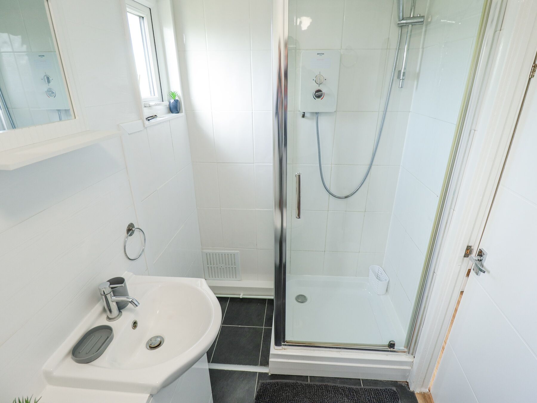 Une salle de bain moderne avec une douche à l’italienne en verre, un lavabo blanc à une seule cuve et une petite fenêtre au-dessus du miroir.