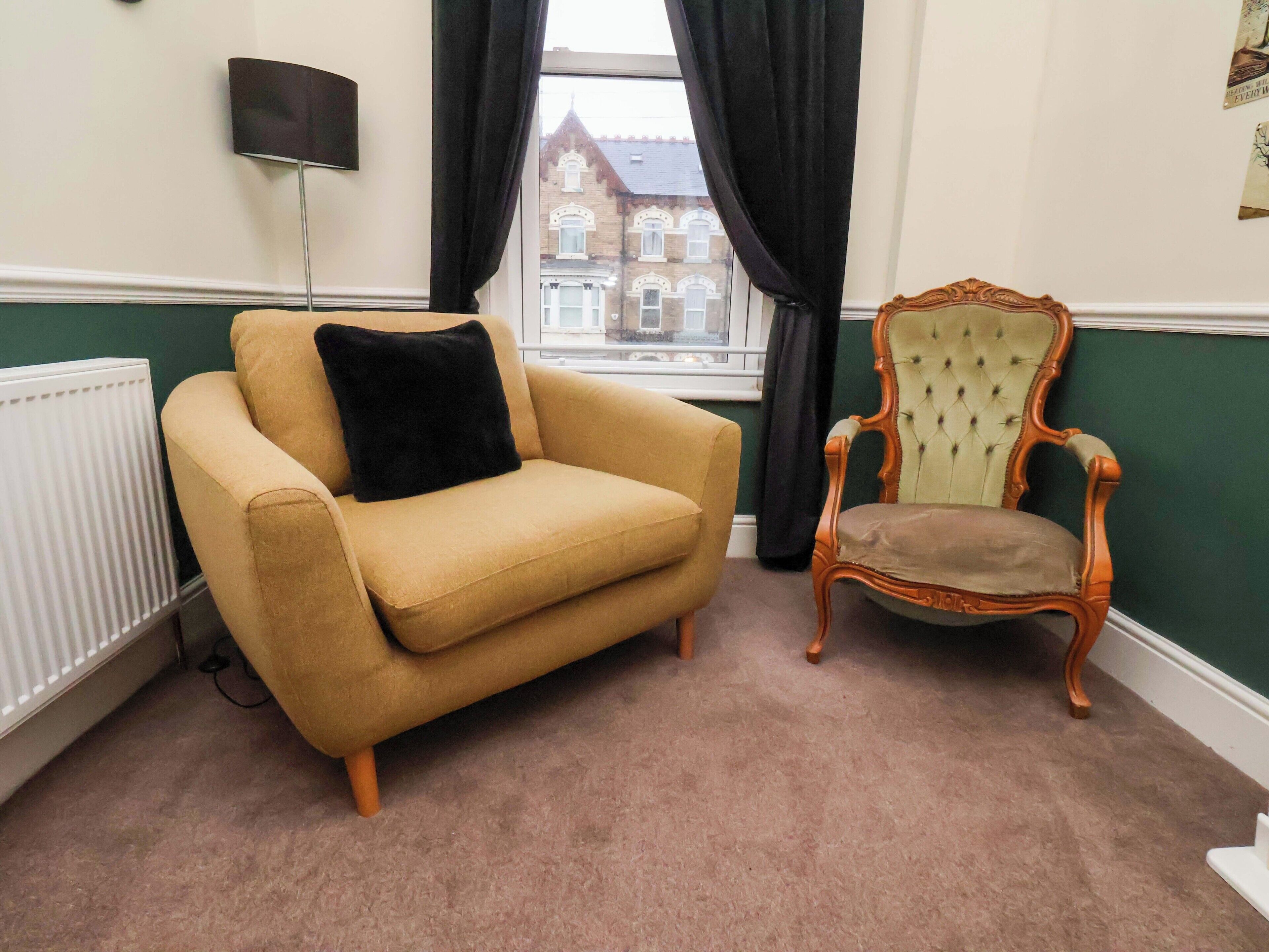 Une pièce comprenant un fauteuil beige, une chaise en bois à dossier capitonné, un radiateur et une fenêtre avec des rideaux.