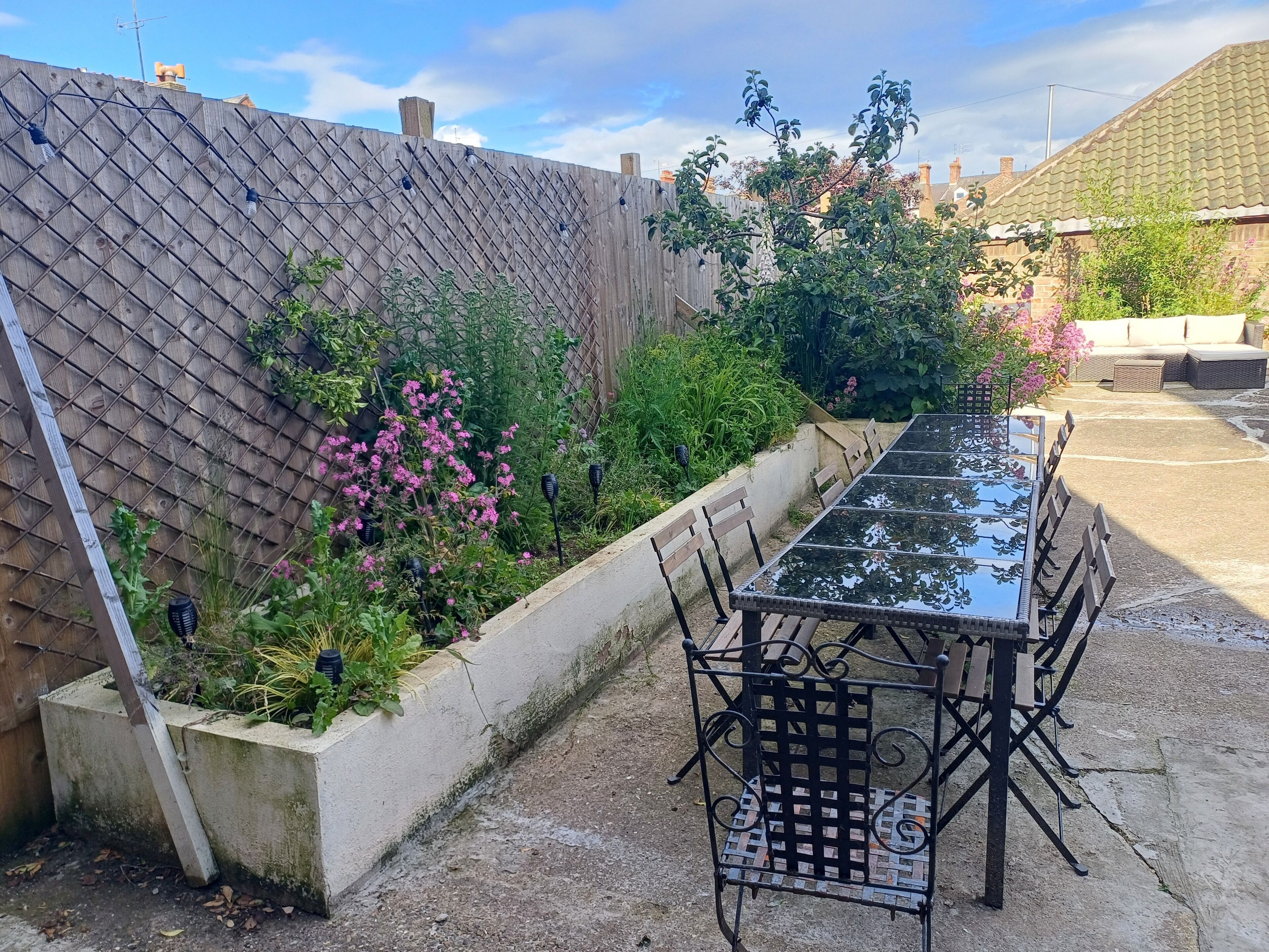 Une terrasse dotée d’une table à plateau de verre et de chaises en métal, entourée d’un jardin où poussent différentes plantes et fleurs.