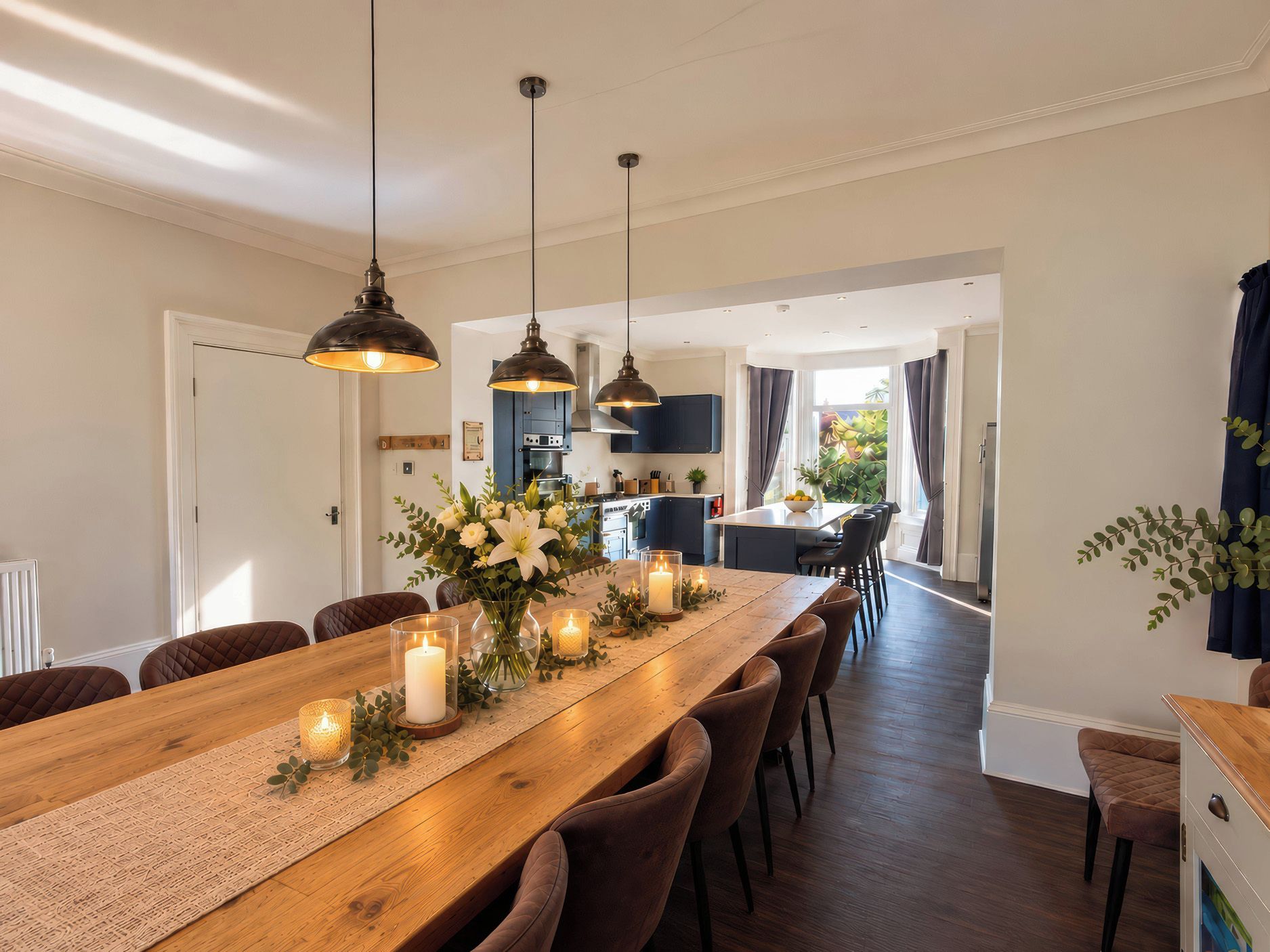 A dining area with a long wooden table, candles, and a vase of flowers. There are chairs around the table, and a kitchen is visible in the background.