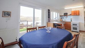 Un comedor con mantel azul, sillas de madera y un centro de mesa con un bol de vidrio. Se ve una cocina con armarios de madera y electrodomésticos blancos al fondo.