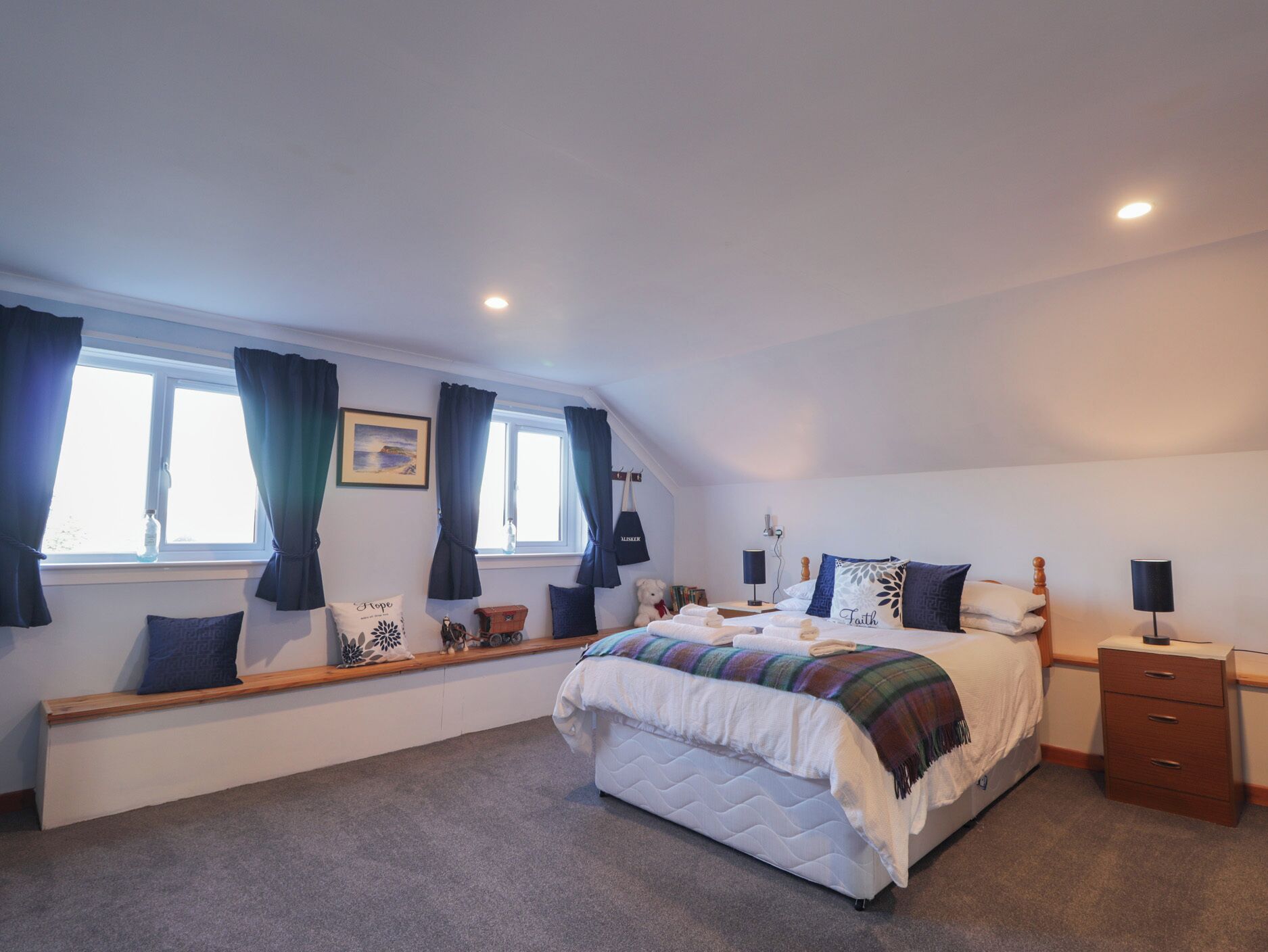 A bedroom with a large bed, a bench with cushions, a window with curtains, and a bedside table with a lamp.