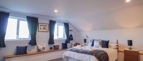 A bedroom with a large bed, a bench with cushions, a window with curtains, and a bedside table with a lamp.