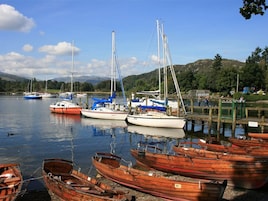 A marina with various boats docked, including sailboats and rowboats, with a backdrop of hills and a clear sky.
