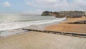 Une plage côtière bordée d’une falaise rocheuse, avec une promenade et des bâtiments en arrière-plan.