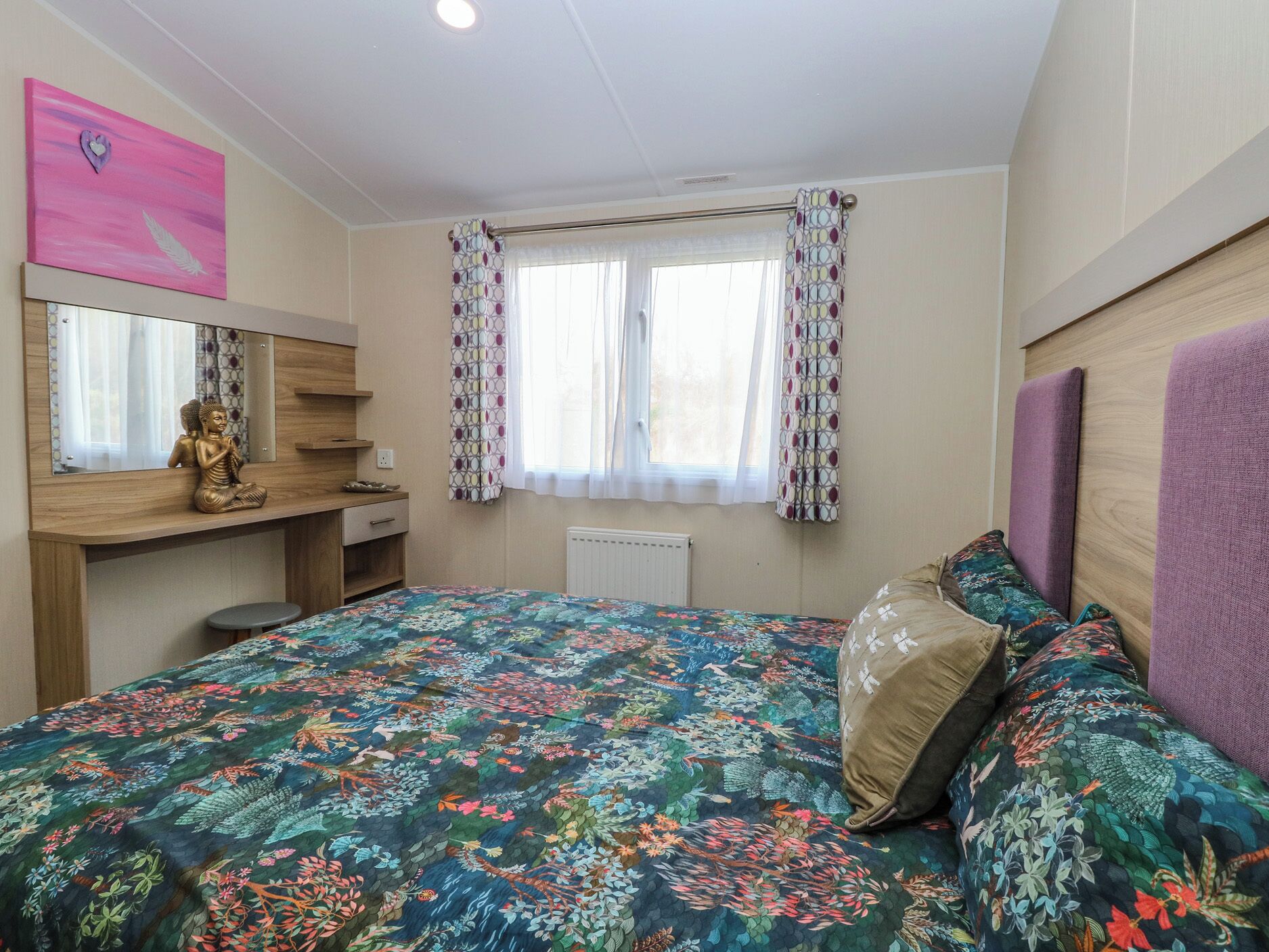 A bedroom with a bed, a desk with a Buddha statue, a mirror, and a window with patterned curtains.