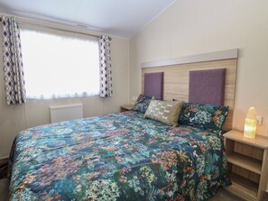 Une chambre à coucher avec une couvre-lit à motifs floraux, deux oreillers, une table de chevet avec une lampe et une fenêtre ornée de rideaux à motifs.