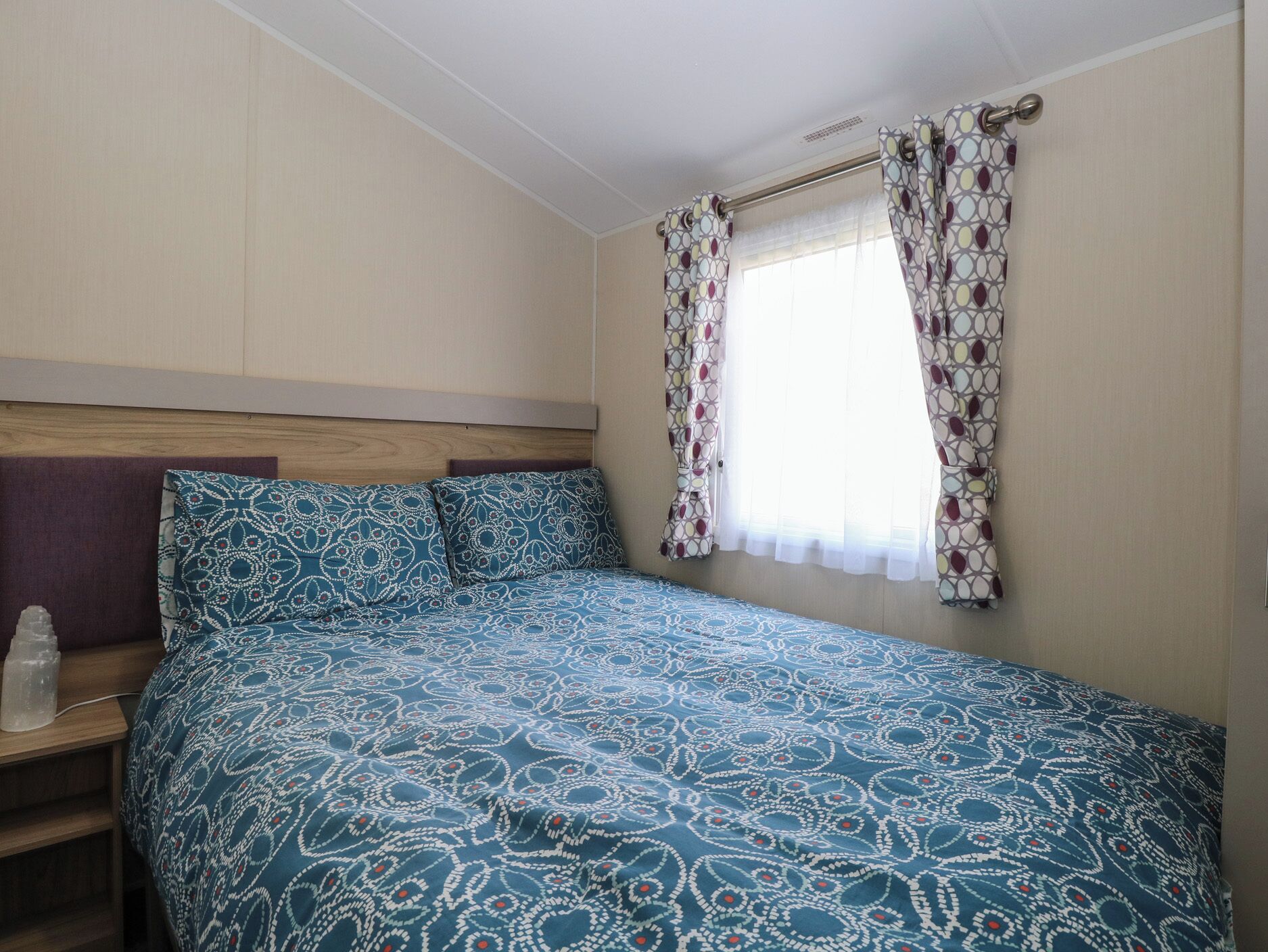 Een slaapkamer met een bed, een raam met gordijnen, een nachtkastje met een fles en een hoofdeinde.