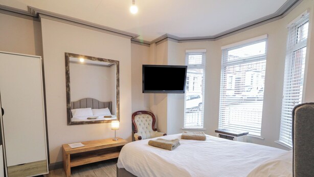 A modern hotel room with a large bed, a flat-screen TV mounted on the wall, a bedside table with a lamp, and a view of the city through the windows. - The Corner House (Londonderry)