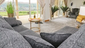 A modern living room with grey sofas, a glass table with breakfast items, and large windows offering a view of greenery and buildings. - Myddelton Grange (Ilkley)