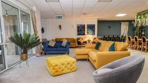 A modern living room with a yellow sofa, a blue sofa, and a grey armchair. There is a yellow ottoman and a potted plant. The room has a large window and a door. - Myddelton Grange (Ilkley)
