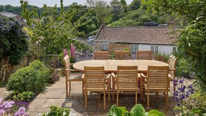 A wooden table and chairs set outdoors with a garden view, stone pathway, and a house with a tiled roof in the background.