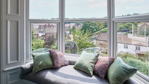 A bay window with a seat and cushions, offering a view of a residential area with houses and greenery.