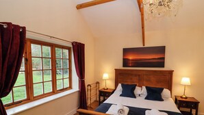 A bedroom with a wooden bed, two bedside tables with lamps, a view of the outdoors through a window, and a painting of a sunset on the wall. - Honeysuckle Cottage (Somerton)