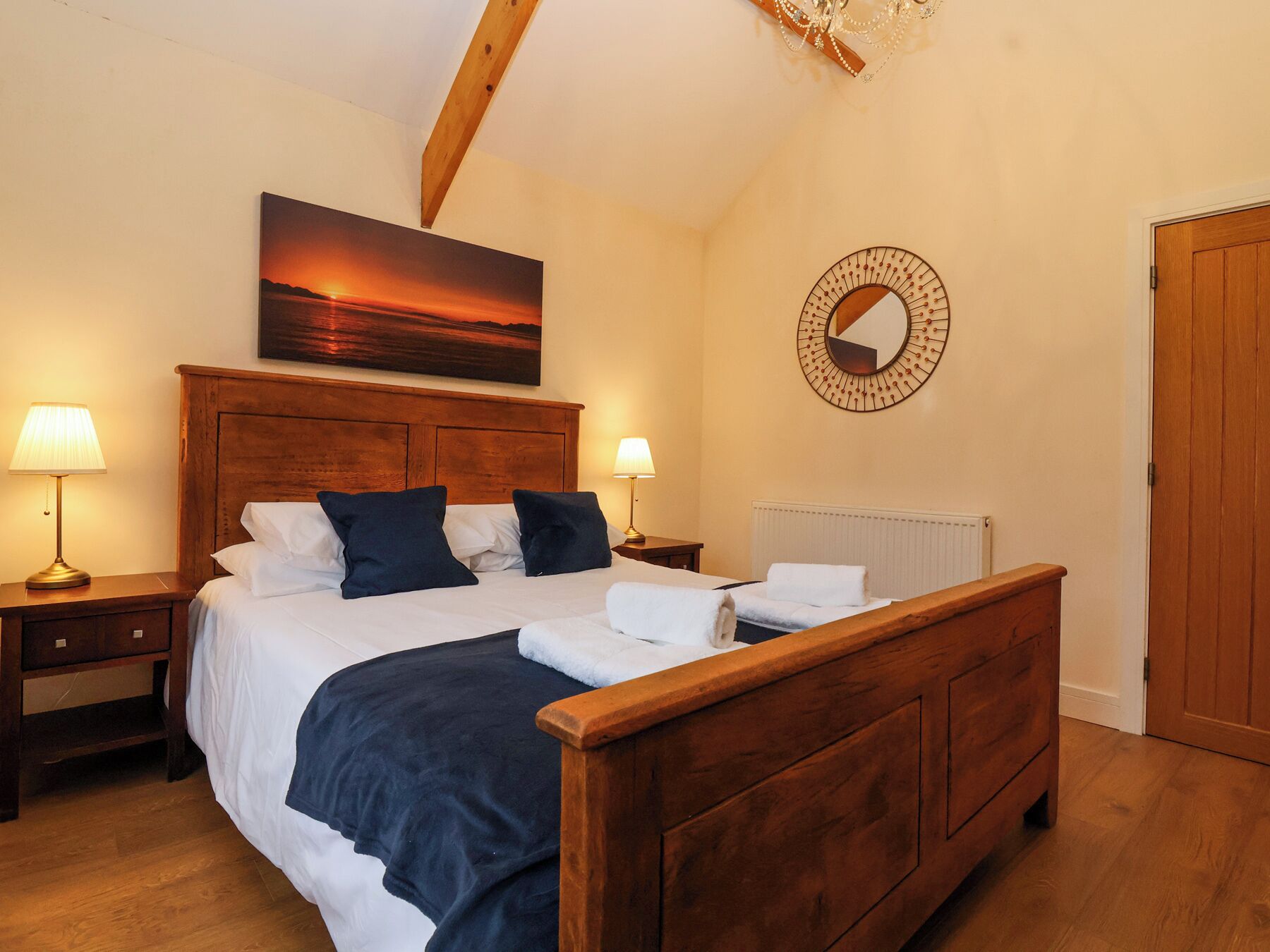 Une chambre avec un lit en bois, deux tables de chevet avec des lampes, un grand tableau représentant un coucher de soleil, un miroir rond et une porte en bois.