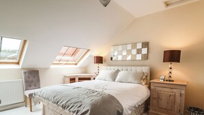 A bedroom with a skylight, a bed with a headboard, bedside lamps, a small table, a chair, and a radiator. - Cob Barn (Exeter)