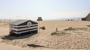 Una casetta sulla spiaggia con tetto a righe e un logo, affacciata su una spiaggia sabbiosa, cielo sereno e montagne sullo sfondo.