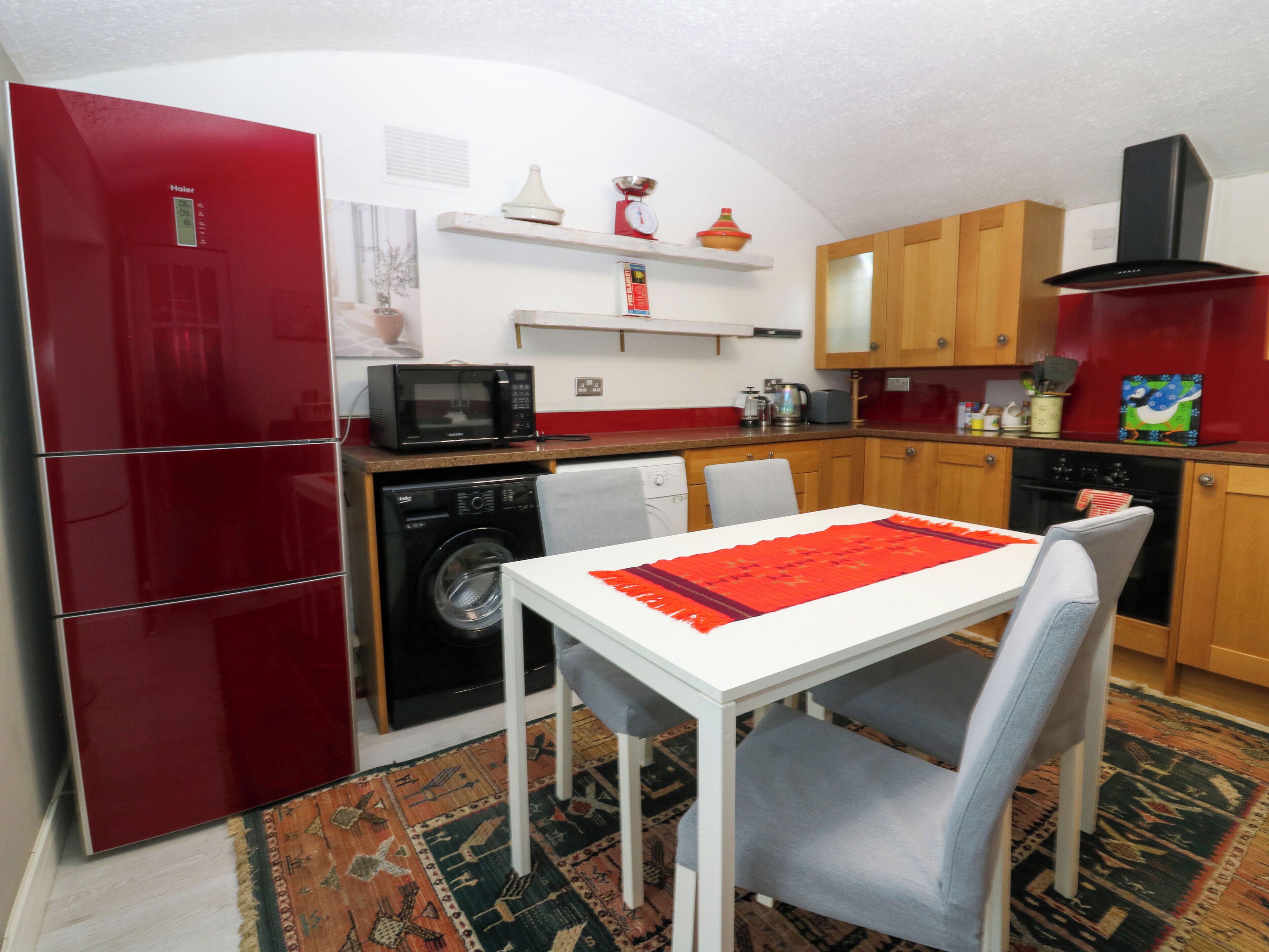 Una cucina compatta con frigorifero rosso, microonde, lavatrice e lavello. C'è un tavolo da pranzo con tovaglia rossa e due sedie. La cucina presenta mobili in legno e un piccolo ripiano con oggetti decorativi.