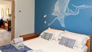 A bedroom with a blue wall, a bed with white and patterned pillows, a rolled towel on the floor, and a white ceiling light. - 3 Palms (Paignton)