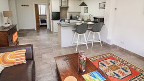 A modern living room with a brown leather sofa, a coffee table, and a colorful rug. A kitchen with a bar area is visible in the background. - 3 Palms (Paignton)