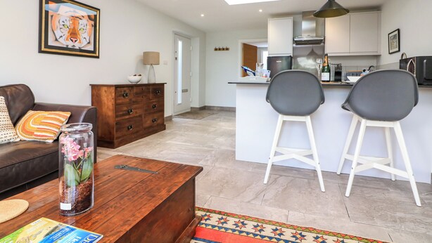 A modern living room with a sofa, coffee table, and a decorative vase. There is a kitchen area with a bar and two chairs. - 3 Palms (Paignton)