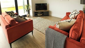 A modern living room with a red sofa, a flat-screen TV, and a large window with a view of a green landscape. - Glynllifon 3 (Marianglas)