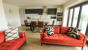 A modern living room with red sofas, a dining area with a table and chairs, and a kitchen with dark cabinets and appliances. - Glynllifon 3 (Marianglas)