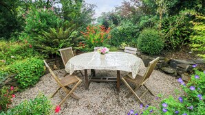 A round table with a floral tablecloth, surrounded by wooden chairs, set on a gravel surface in a garden with various plants and flowers. - 2 Summerhill (Haverfordwest)