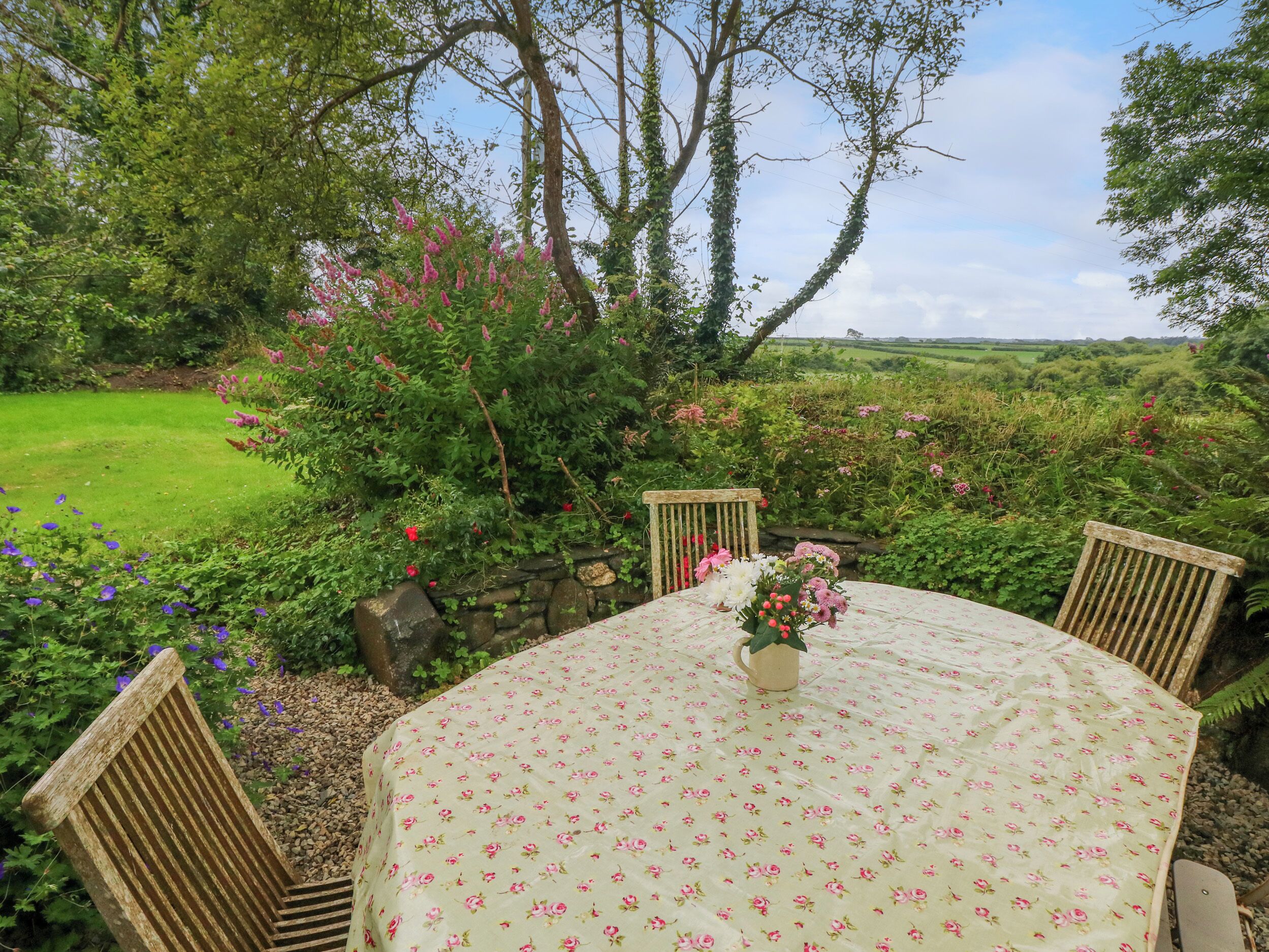 A round table with a floral tablecloth, surrounded by wooden chairs, set in a lush garden with a stone wall and various plants.
