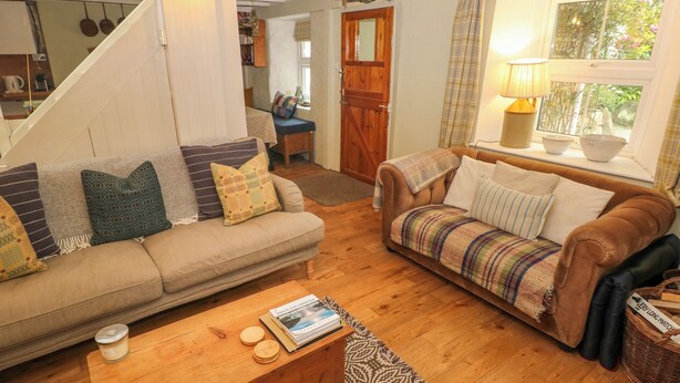 A cozy living room with a sofa, a coffee table, and a lamp. There is a window with curtains and a view of greenery outside. - 2 Summerhill (Haverfordwest)